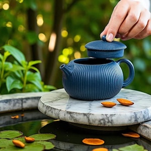 以下が含まれることがあります： ネイビーブルーのセラミックティーポット、リブのデザイン。蓋が持ち上げられ、内部が明らかになります。ティーポットは、スイレンと浮遊する種子のさやがある水の近くの大理石の台の上にあります。