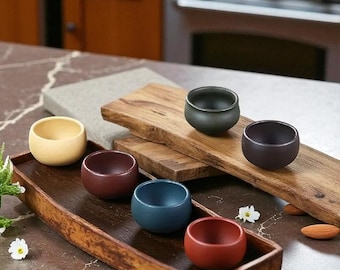 May include: A collection of small, round ceramic bowls in various colors, including yellow, burgundy, and blue. The bowls are arranged on a wooden tray and a wooden plank, suggesting a serving or display setting. The background features a kitchen setting.