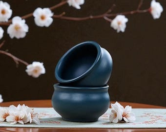 May include: Two stacked, teal-colored ceramic bowls. The top bowl is tilted, revealing its interior. The bowls sit on a patterned surface, with white flowers and branches in the background. The bowls have a smooth, matte finish.