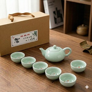May include: A pale green ceramic tea set comprising a teapot and six small cups, each featuring a fish design. The set is presented on a wooden surface, alongside a brown gift box with a decorative label.