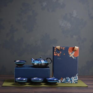 May include: A ceramic tea set in blue and black, comprising a teapot and four cups. The teapot and cups have a speckled pattern. The set is presented on a blue box and a green mat, with a gift box featuring a crane illustration.
