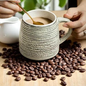 May include: A ceramic coffee mug with a textured, striped design, filled with coffee and stirred with a gold spoon. The mug sits on a bed of coffee beans, with a small white creamer pitcher in the background. The mug has a large handle.