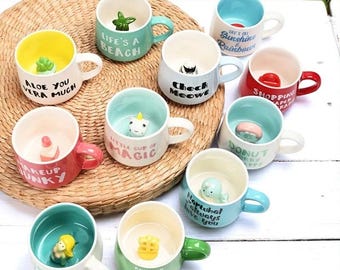 Puede incluir: Una colección de tazas de cerámica con varios diseños y colores. Cada taza presenta una figura interior única y una frase, como "Life's a Beach" o "Check Meow". Las tazas tienen asas e interiores de diferentes colores, dispuestas sobre una estera tejida.