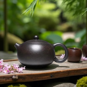 Könnte beinhalten: Eine dunkelbraune Keramik-Teekanne mit rundem Körper, gebogenem Henkel und kleinem Ausguss. Die Teekanne steht auf einer Holzoberfläche mit zwei kleinen braunen Teetassen und rosa Blüten. Der Hintergrund ist ein verschwommener grüner Garten.