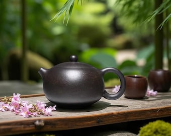 May include: A dark brown ceramic teapot with a rounded body, a curved handle, and a small spout. The teapot sits on a wooden surface with two small brown teacups and pink flowers. The background is a blurred green garden.