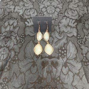 May include: A pair of gold-toned drop earrings with two light green, teardrop-shaped stones. The earrings are displayed on a black card against a dark gray floral patterned background. The earrings have a simple, elegant design.
