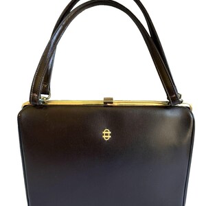 Puede incluir: Un bolso vintage marrón oscuro con dos asas curvas. El bolso rectangular tiene un cierre dorado y un pequeño emblema dorado en la parte delantera. Las asas también son de color marrón oscuro.