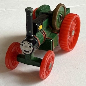 May include: A green and black toy steam engine with red wheels. The engine features a smiling face and a black smokestack. The wheels are a vibrant red. This miniature replica of a classic steam engine is a charming toy.