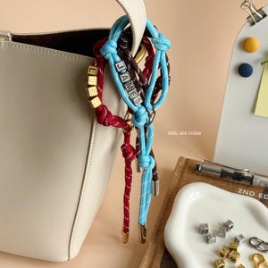 Könnte beinhalten: Cremefarbene Handtasche mit weißem Henkel, verziert mit farbigen Seilarmbändern. Ein Armband schreibt "JAMES" in silbernen Buchstaben, ein anderes "LISA" in Gold. Die Armbänder sind rot, blau und braun.