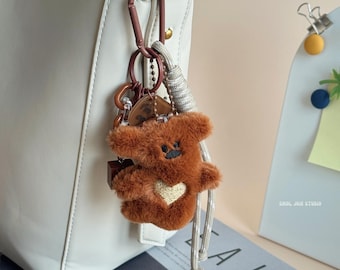 May include: A brown teddy bear keychain with a gold heart on its chest, attached to a white bag with a brown carabiner clip. The keychain also includes a small wooden box and other charms. The bear has a black embroidered face.