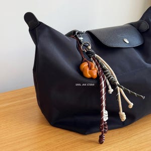 Pode incluir: Uma bolsa tote preta com um chaveiro de corda marrom e branco. O chaveiro tem um pequeno nó marrom e o texto "SEOL JOIE STUDIO" está impresso na bolsa.