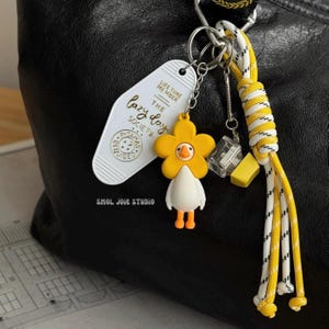 May include: A keychain with a white hotel tag reading "lazy day society" and a yellow flower duck charm. The keychain also has a clear cube, a yellow block, and a yellow and white rope lanyard. The overall design is bright and playful.