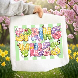 以下が含まれることがあります： 「SPRING VIBES」の文字がカラフルなパッチワーク風の文字で描かれた白いトートバッグ。文字は花と蝶で装飾され、緑と白のストライプの背景に配置されています。バッグは春の庭を背景に掲げられています。