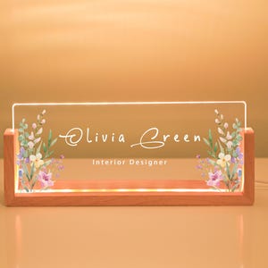 May include: A desk nameplate with the name "Olivia Green" and the title "Interior Designer" in white script. The clear acrylic plate is set in a wooden base and decorated with watercolour floral designs.