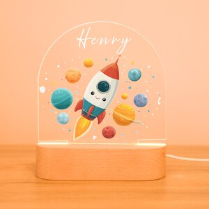 Peut inclure: Veilleuse décorative avec une fusée de dessin animé souriante, entourée de planètes. Le nom "Henry" est écrit au-dessus de la fusée. La lumière repose sur une base en bois et est alimentée par un cordon.
