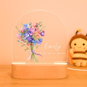 May include: A night light with a wooden base and a clear acrylic panel. The panel displays a watercolour bouquet of purple, blue, pink, and orange flowers, with the name "Emily" and text below. A plush bee toy is in the background.