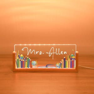 May include: A wooden-framed LED light with the name "Mrs. Allen" in script. The light features a colourful illustration of pencils, globes, and books, perfect for a teacher's desk. The light is plugged into an electrical outlet.