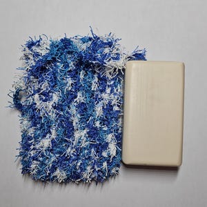 May include: A white bar of soap sits next to a blue and white crocheted soap sack.