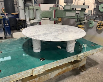 May include: A round marble table with a white and gray pattern, supported by three cylindrical legs. The table is set on a large, green industrial surface, suggesting a workshop setting. The table's diameter is approximately 48 inches.