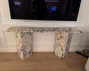 May include: A marble console table with a rectangular top and two geometric legs. The table features a white and gray veined pattern. The table is placed against a white wall and a wooden floor.