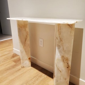May include: White marble console table with two thick, rectangular legs. The table has a white top and a natural marble pattern on the legs.