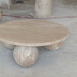 May include: A round, light brown stone coffee table with a smooth surface. The table is supported by three spherical stone bases. Another similar table is visible in the background.
