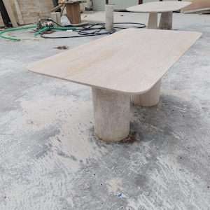 May include: A rectangular travertine table with two cylindrical legs. The table is made of natural stone and has a light beige color.