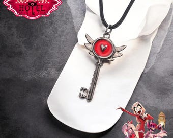 May include: A silver key necklace with red wings and a heart-shaped charm. The key has a red heart in the centre of the charm. The necklace is on a black cord. The text "HAZBIN HOTEL" is visible in the background.