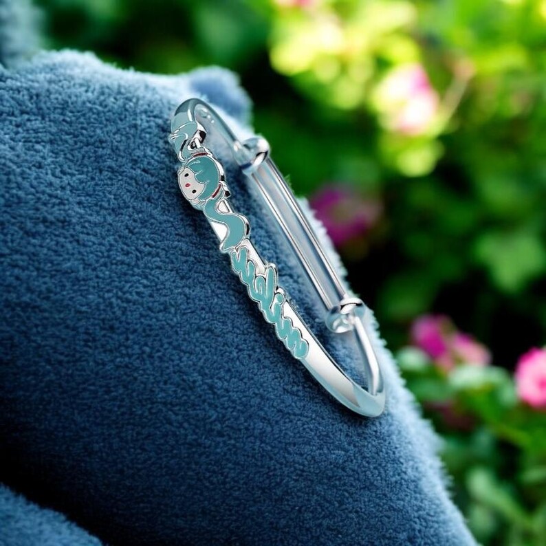 Pu&ograve; includere: Un braccialetto rigido d'argento con un design azzurro e bianco. Il braccialetto presenta un personaggio dei cartoni animati e la parola "Sunshine" in azzurro. Il braccialetto &egrave; appoggiato su una superficie blu.