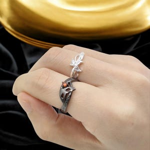 May include: Two rings are displayed on a hand. One is silver with a butterfly design. The other is black with a mask design and a small orange stone. The rings are on a hand with a gold and black background.