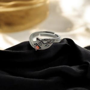 May include: A silver-colored adjustable ring featuring a fox head design with cut-out details and a small, round, orange gemstone. The ring is set against a black fabric background.