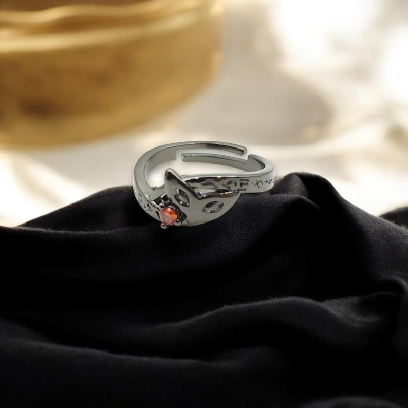 May include: A silver-colored adjustable ring featuring a fox head design with cut-out details and a small, round, orange gemstone. The ring is set against a black fabric background.