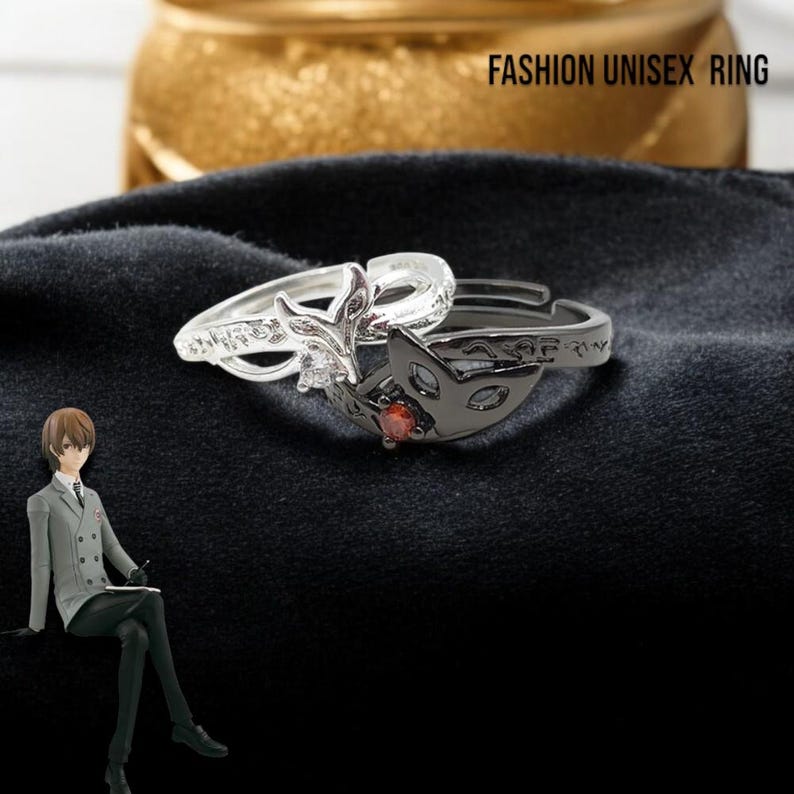 May include: Two rings, one silver and one black, are displayed. The silver ring features a floral design with a central clear stone. The black ring has a mask design with a red stone. The text "FASHION UNISEX RING" is visible.
