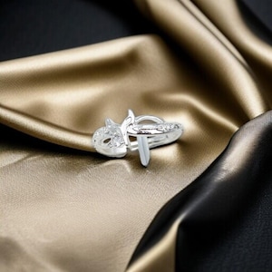 May include: A silver ring with a unique design featuring a stylized cat face with clear gemstones and a vertical bar. The ring is set against a backdrop of gold and black fabric.