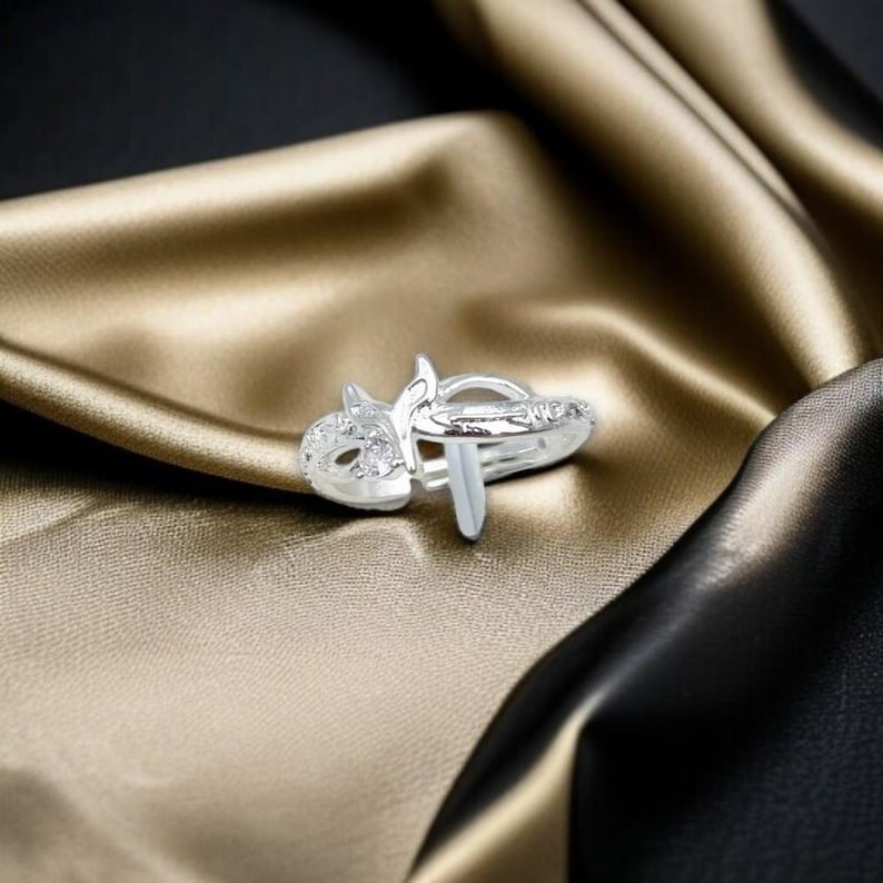 May include: A silver ring with a unique design featuring a stylized cat face with clear gemstones and a vertical bar. The ring is set against a backdrop of gold and black fabric.