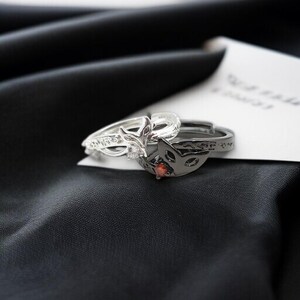 May include: Two rings, one silver and one black, rest on a black satin surface. The silver ring features a fox head design with a clear stone. The black ring has a fox mask design with a red stone.