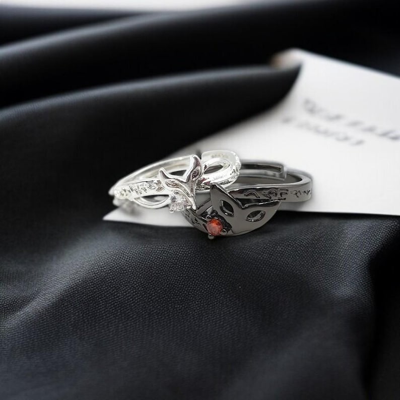 May include: Two rings, one silver and one black, rest on a black satin surface. The silver ring features a fox head design with a clear stone. The black ring has a fox mask design with a red stone.