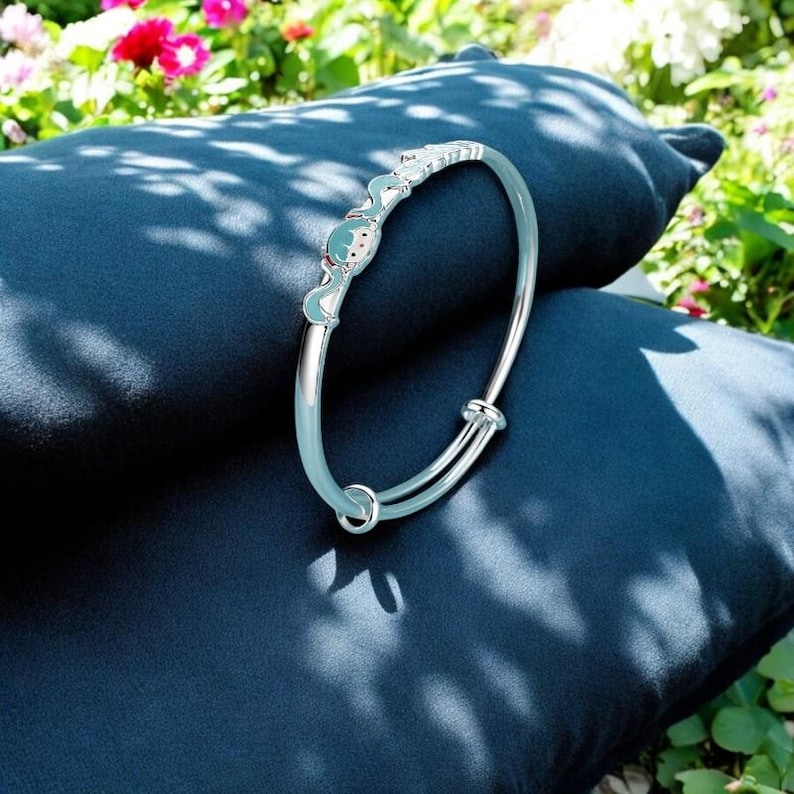 Pu&ograve; includere: Un braccialetto regolabile color argento con rivestimento in smalto azzurro. Il braccialetto presenta una sezione decorativa con un design di personaggi dei cartoni animati in azzurro, bianco e rosso. Il braccialetto &egrave; appoggiato su una superficie blu scuro.