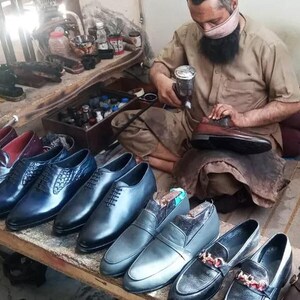 Puede incluir: Una variedad de zapatos de vestir para hombre en cuero azul, marr&oacute;n y negro. Los zapatos est&aacute;n dispuestos sobre una mesa de madera. Un hombre est&aacute; trabajando en un zapato en el fondo.