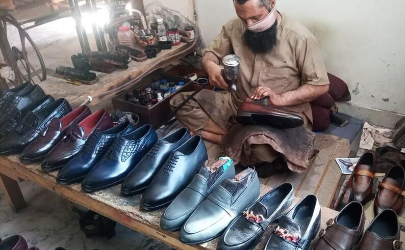 Puede incluir: Una variedad de zapatos de vestir para hombre en cuero azul, marr&oacute;n y negro. Los zapatos est&aacute;n dispuestos sobre una mesa de madera. Un hombre est&aacute; trabajando en un zapato en el fondo.