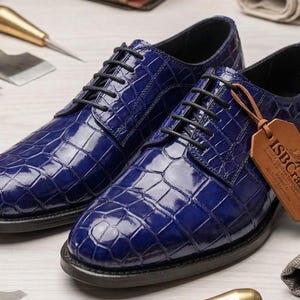May include: A pair of glossy royal blue oxford shoes featuring a textured crocodile skin pattern, black laces, and a dark sole. A tan leather tag with the embossed text ISBCrafted is attached to the right shoe, which has a rounded toe design.