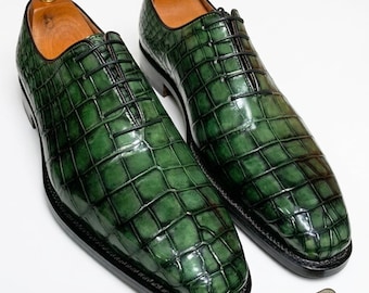 May include: A pair of emerald green oxford shoes featuring a glossy crocodile skin texture, black laces, and wooden shoe trees. A rectangular brown leather tag with the text ISBCrafted Est. 1995 is attached by a thin brown cord.