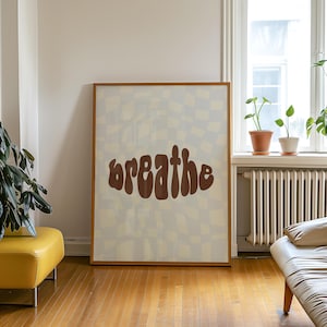 Könnte beinhalten: Ein gerahmter Kunstdruck mit dem Wort "breathe" in braunen, stilisierten Buchstaben. Der Hintergrund hat ein wellenförmiges Karomuster in Creme und Hellbraun. Der Rahmen ist hellbraun. Der Druck wird in einem Raum mit einem gelben Hocker und Pflanzen ausgestellt.