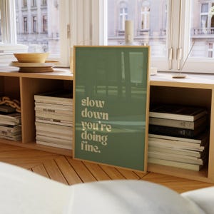 May include: A framed art print with the text "slow down you're doing fine" in a serif font. The background is a solid muted green color. The frame is a light wood color. The print is displayed on a wooden shelf.