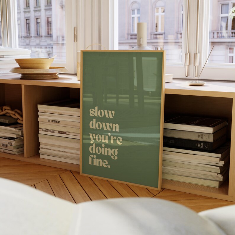 May include: A framed art print with the text "slow down you're doing fine" in a serif font. The background is a solid muted green color. The frame is a light wood color. The print is displayed on a wooden shelf.