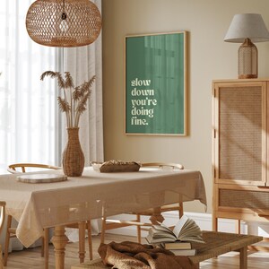May include: A dining room scene with a beige tablecloth, wooden chairs, and a woven pendant light. A framed green poster reads "slow down you're doing fine." A vase holds dried plants. A bench holds books and a brown throw.