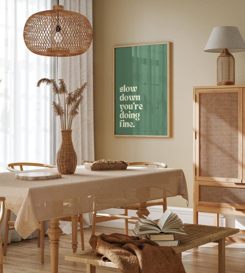 May include: A dining room scene with a beige tablecloth, wooden chairs, and a woven pendant light. A framed green poster reads "slow down you're doing fine." A vase holds dried plants. A bench holds books and a brown throw.