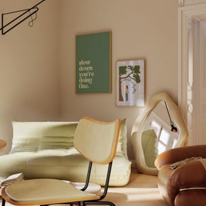 May include: A modern living room scene featuring a light green sofa, a wooden chair, and two framed art prints. One print reads "slow down you're doing fine." A large, abstract mirror and a brown leather armchair complete the space.