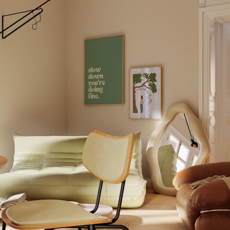 May include: A modern living room scene featuring a light green sofa, a wooden chair, and two framed art prints. One print reads "slow down you're doing fine." A large, abstract mirror and a brown leather armchair complete the space.
