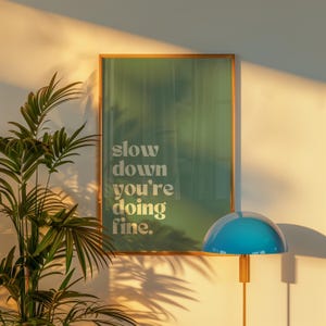 May include: A framed poster with the text "slow down you're doing fine" in white lettering. The poster has a green background and is hanging on a white wall. A blue lamp with a gold stand is in the foreground.